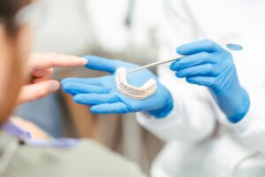 A dentist in blue gloves holds a dental mold with one hand and points with a dental tool, while a patient’s hand gestures toward the mold. The scene suggests a dental consultation or treatment explanation.