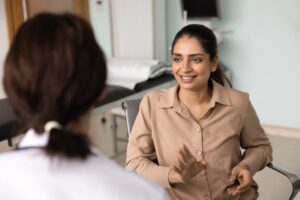 A woman in a tan shirt sits in a medical office, smiling and gesturing as she talks to a healthcare professional in a white coat whose back is to the camera. Medical equipment can be seen in the background.